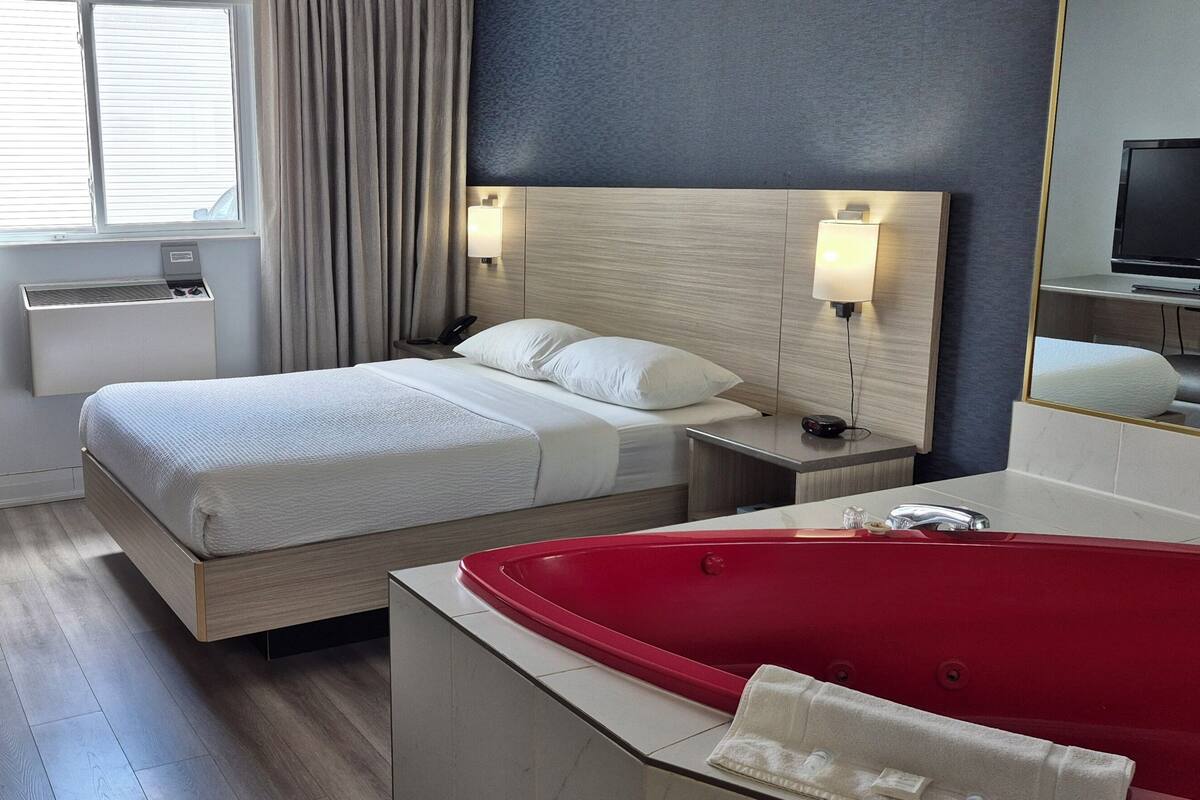 A hotel room with a bed, bedside tables, a red bathtub, a TV, and a window with curtains.
