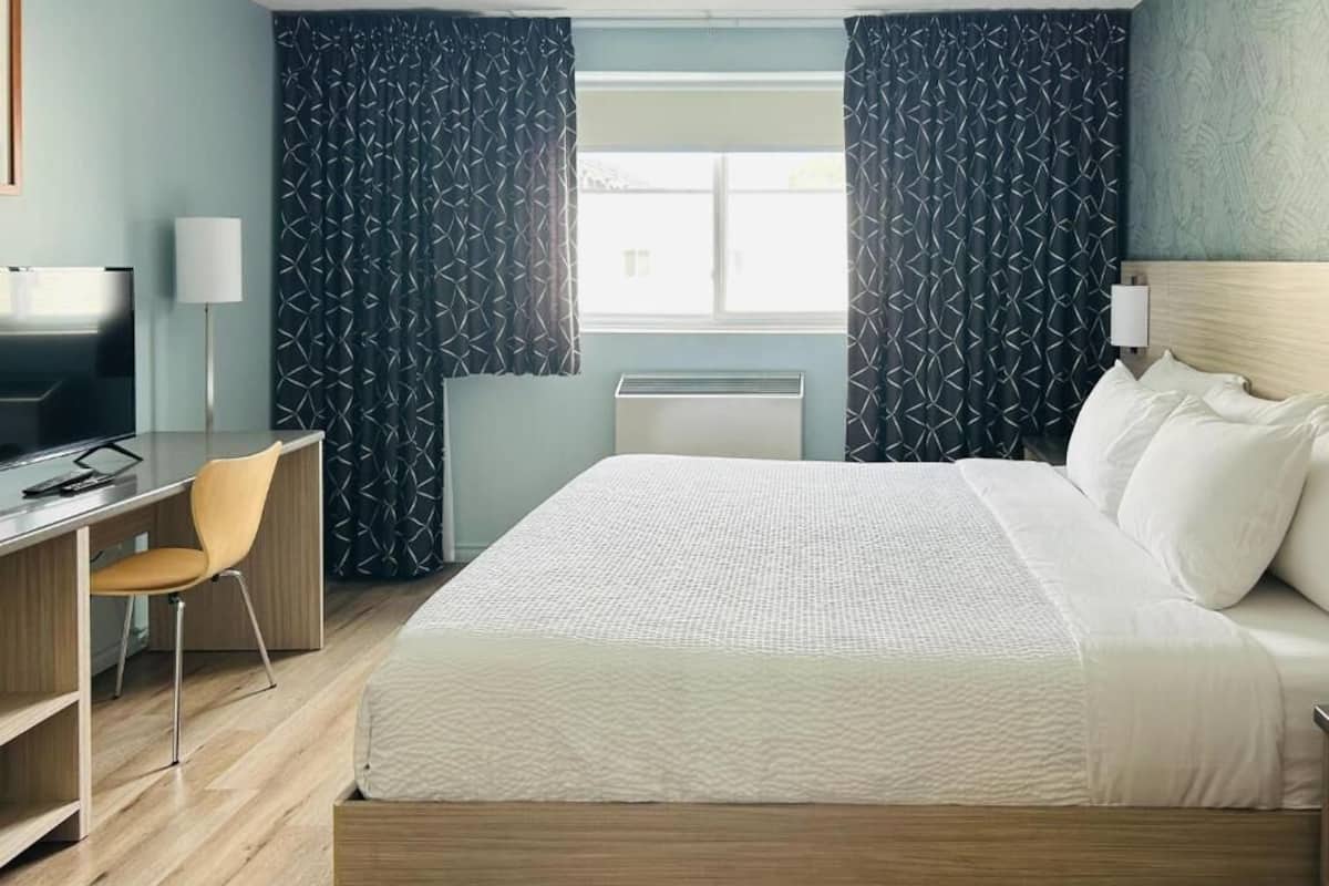 A hotel room with a bed, a desk with a chair, a television, and a window with curtains.