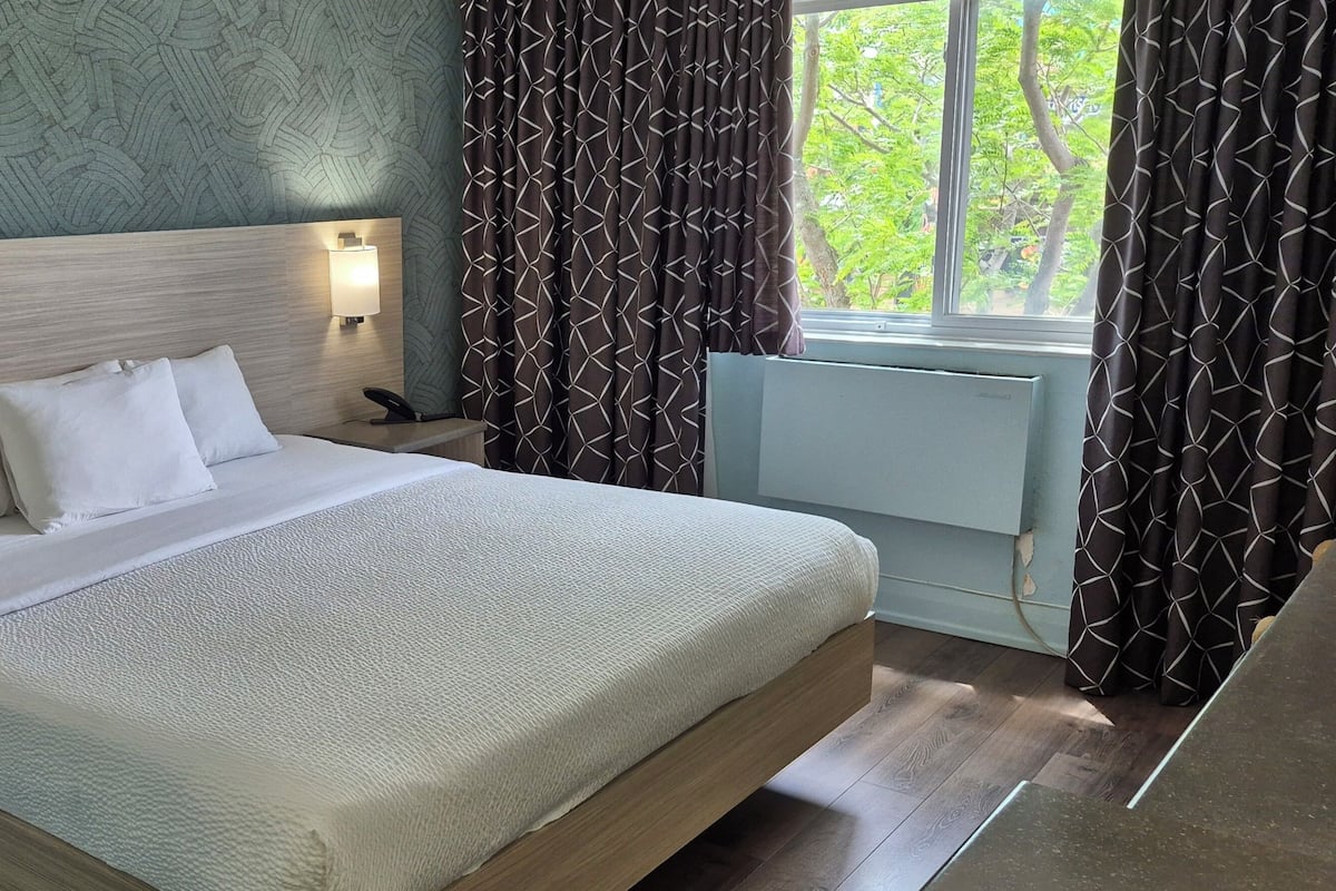 A neatly made bed with white linens, a wooden headboard, bedside lamps, and a window with curtains.