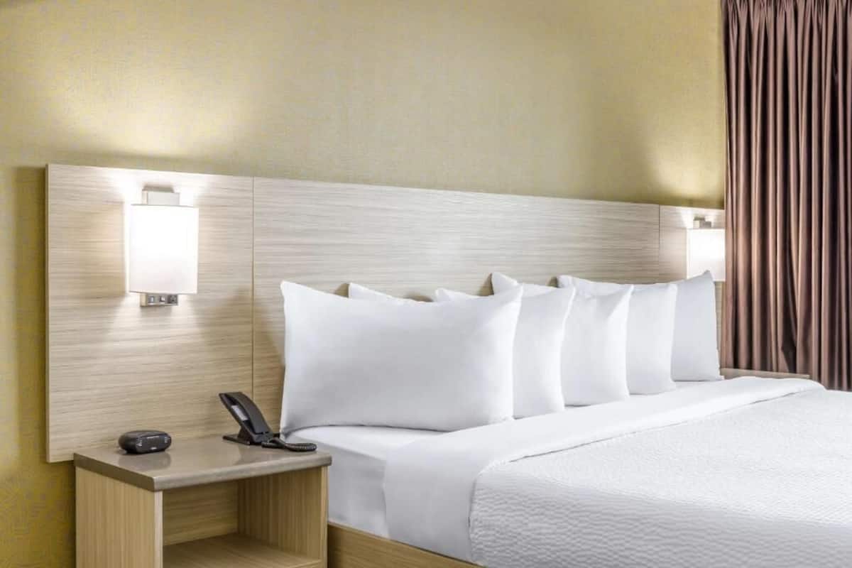 A hotel room with a wooden headboard, white bedding, a bedside table with a phone, and a lamp.