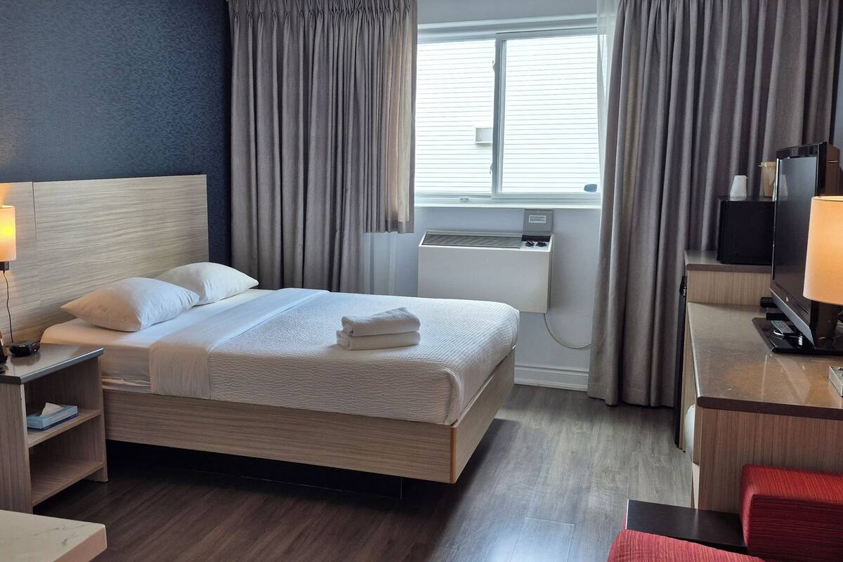 A hotel room with a bed, bedside table, lamp, TV, and a desk with a chair.