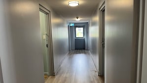 A hallway with light-colored walls and wooden flooring, leading to a door with an exit sign. - Mammas Inn (Niagara Falls)