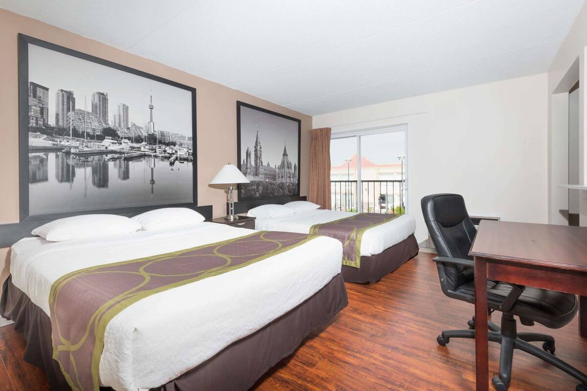 Habitación de hotel con dos camas, un escritorio, una silla y ventanas amplias con vistas a un paisaje urbano.