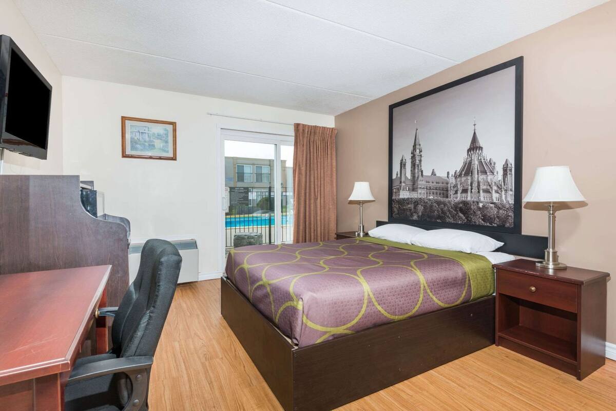 Una habitación de hotel con cama, escritorio, silla, dos lámparas, televisor y un cuadro grande en la pared.
