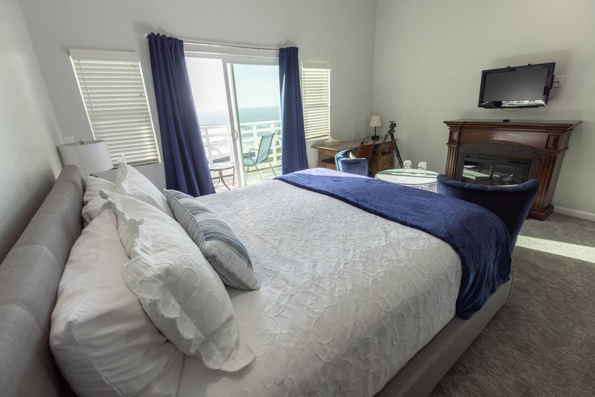 A hotel room with a large bed, a TV mounted on the wall, a fireplace, and a view of the ocean through the windows.