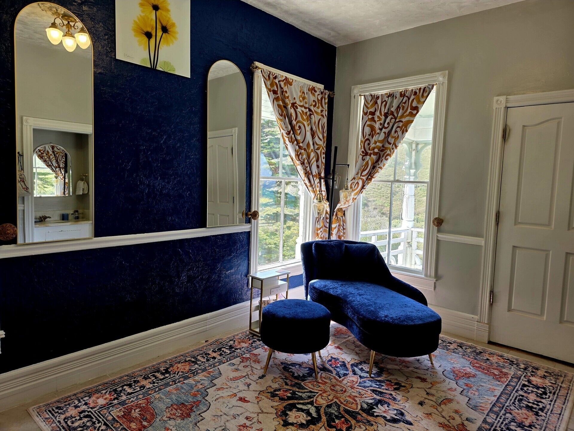 Una habitación con pared azul oscuro, un espejo grande, un sillón azul, un tapete estampado y una ventana con cortinas.