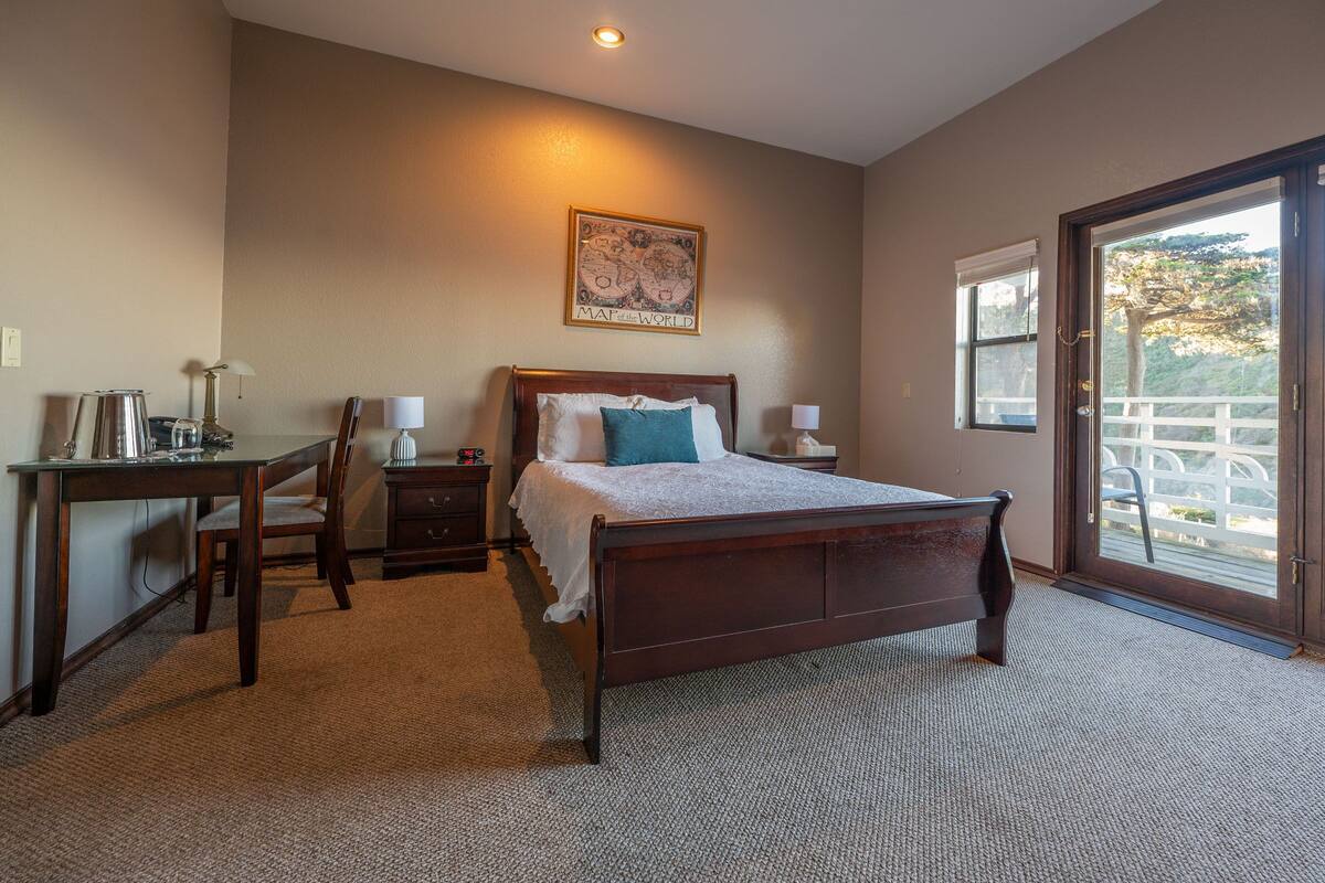 A bedroom with a wooden bed, a desk, a chair, a nightstand, and a window with a view.