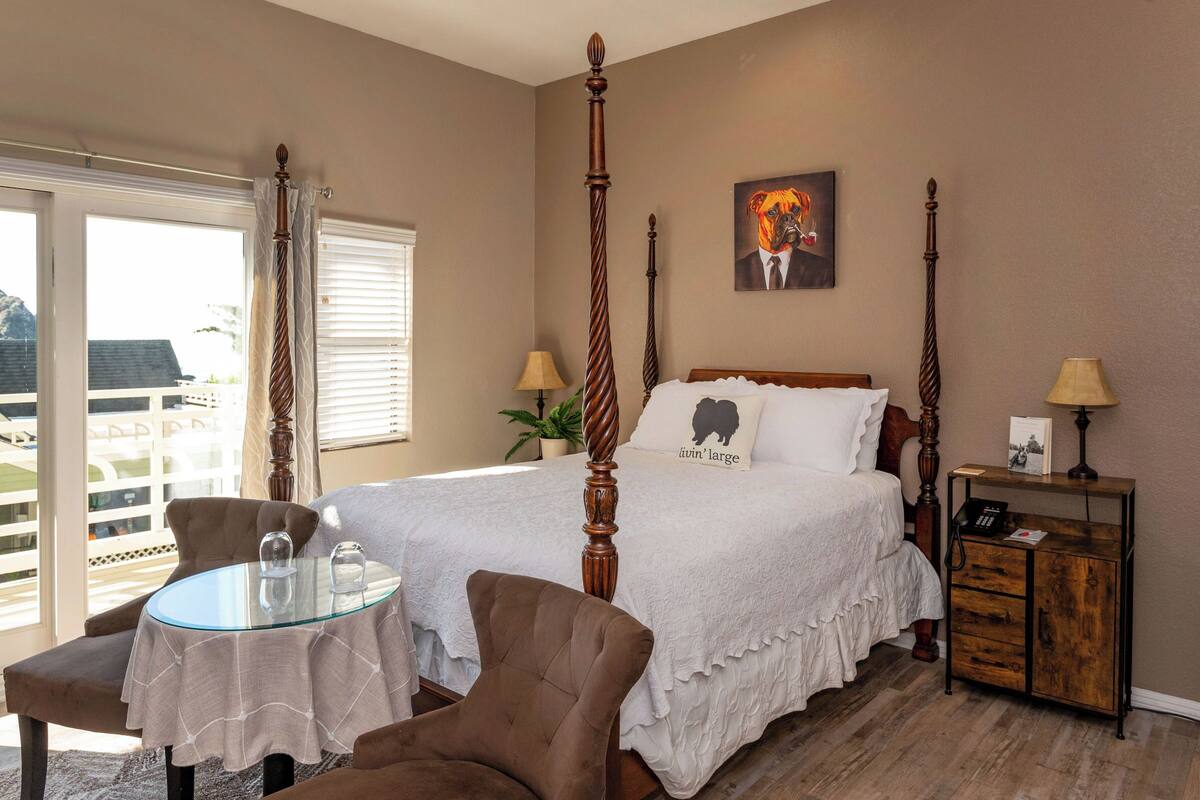 A bedroom with a four-poster bed, a glass table, a chair, a bedside table with a lamp, and a window with blinds.