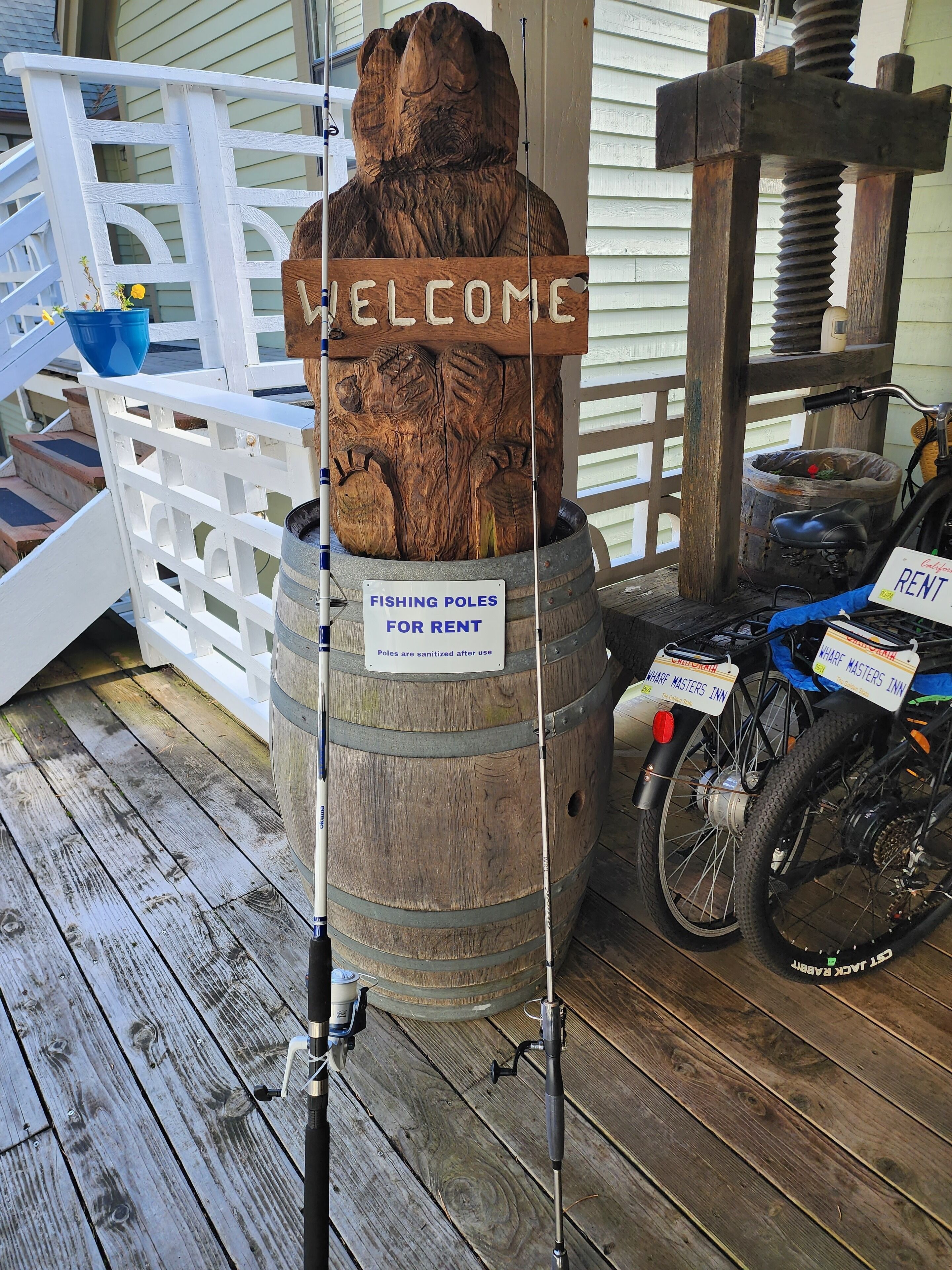 A wooden barrel with a welcome sign, a sign indicating fishing poles for rent, and bicycles with rental tags.