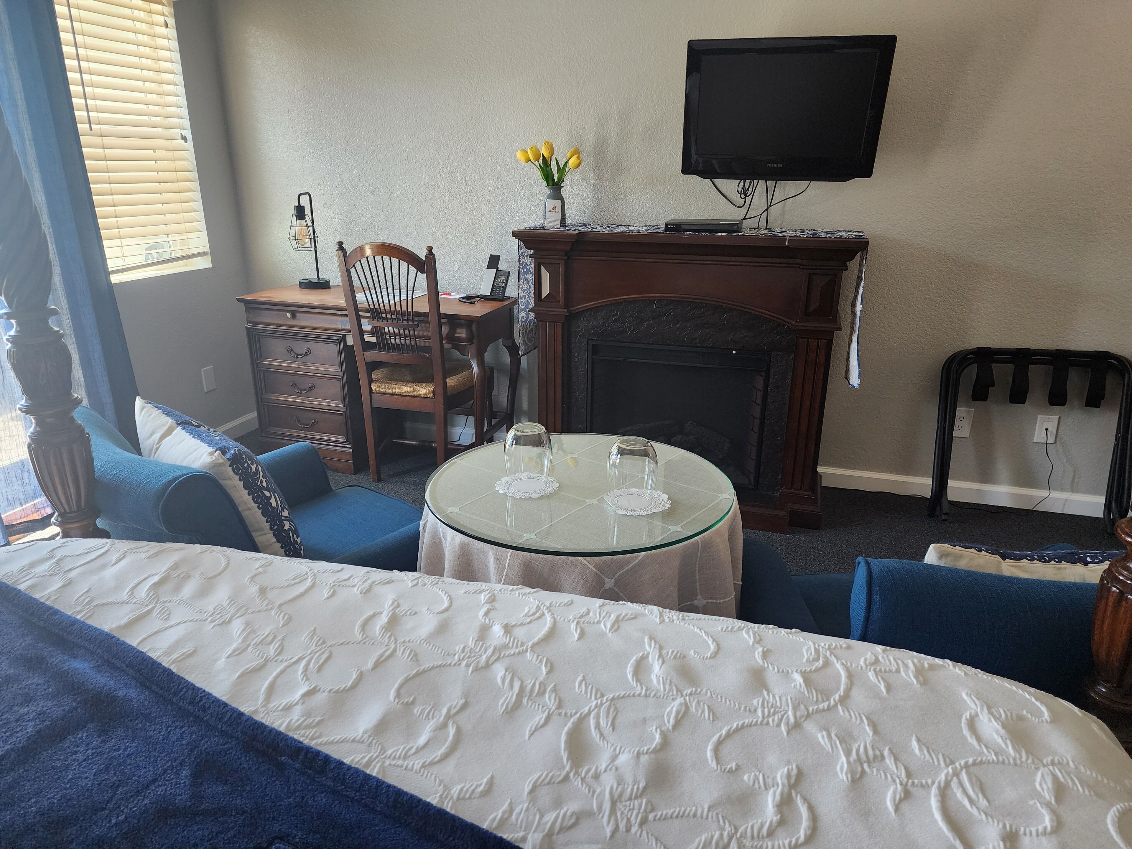 Habitación de hotel con una cama, una mesa redonda de cristal con mantel blanco, una silla de madera, chimenea y un televisor de pantalla plana montado en la pared.