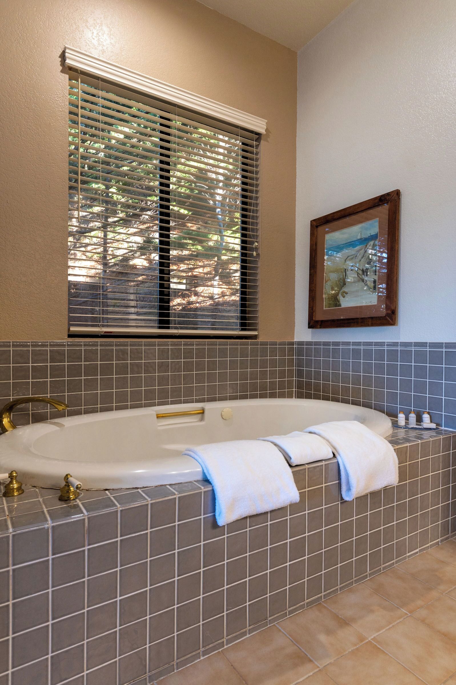A bathroom with a large bathtub, tiled walls, a window with blinds, and a framed picture on the wall.
