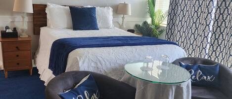 Une chambre d’hôtel avec un lit, deux lampes de chevet, un tableau, une table en verre, deux fauteuils et un tapis bleu.
