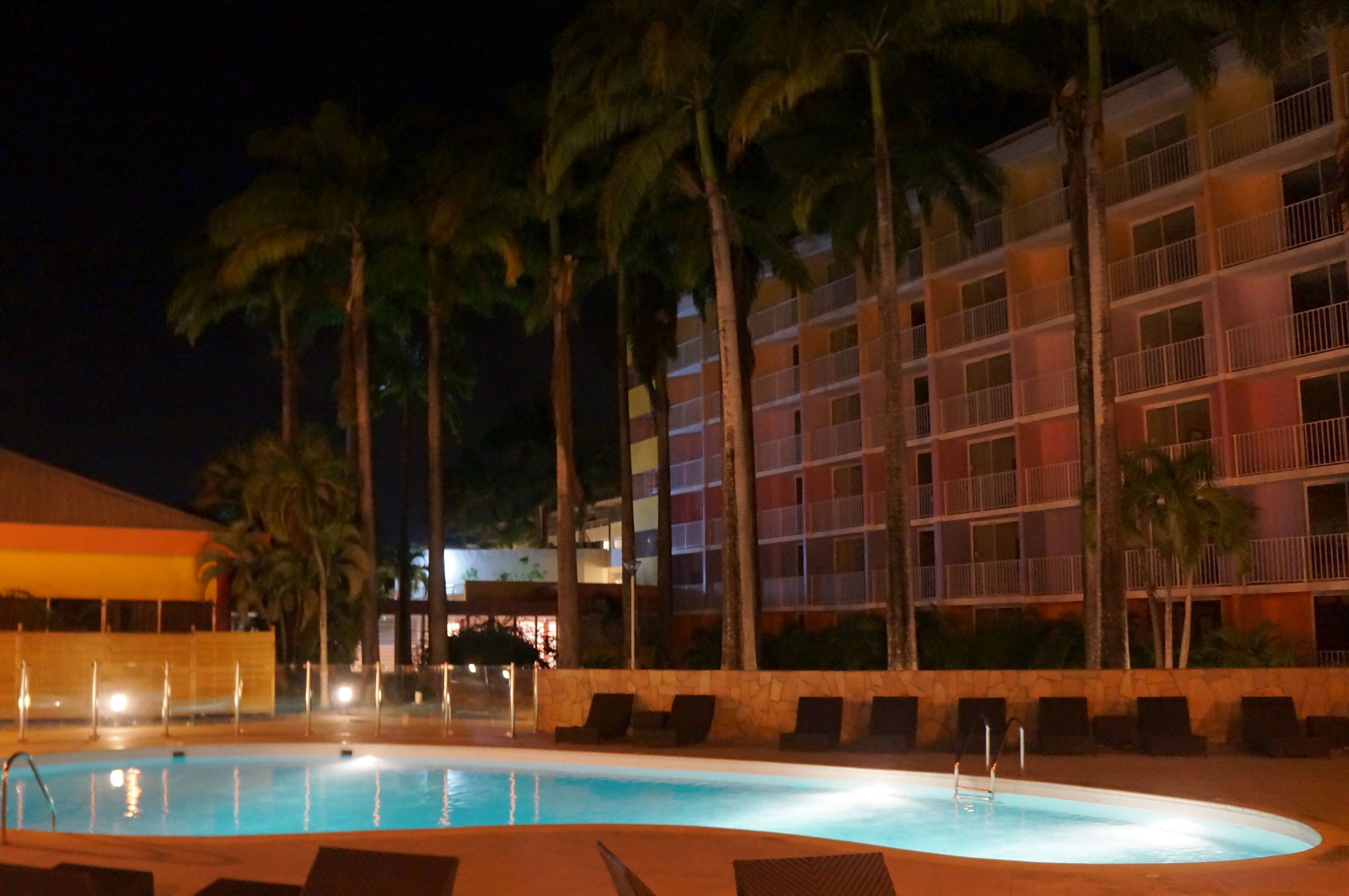 Una piscina rodeada de sillas de descanso y palmeras, con un edificio de varios pisos al fondo.