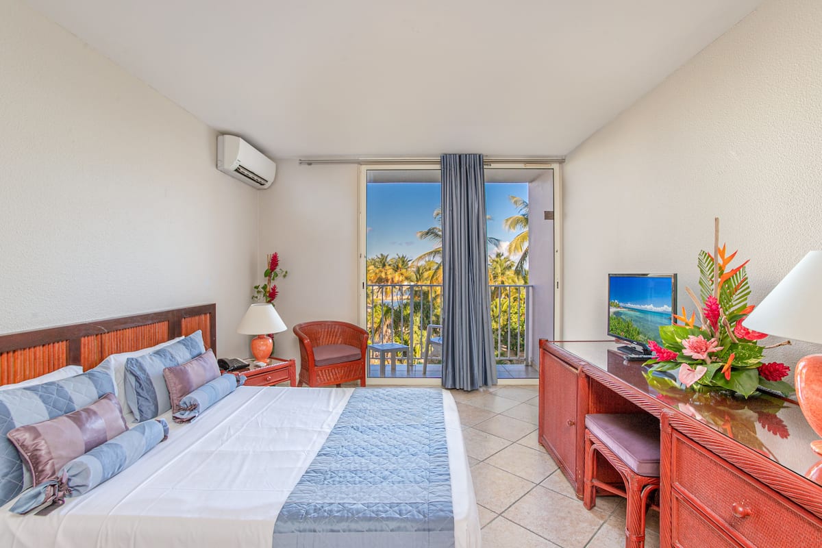 Een hotelkamer met een bed, bureau, stoel, televisie en balkon met uitzicht op palmbomen.