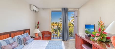 Habitación de hotel con cama, escritorio, silla, televisión y balcón con vista a palmeras.