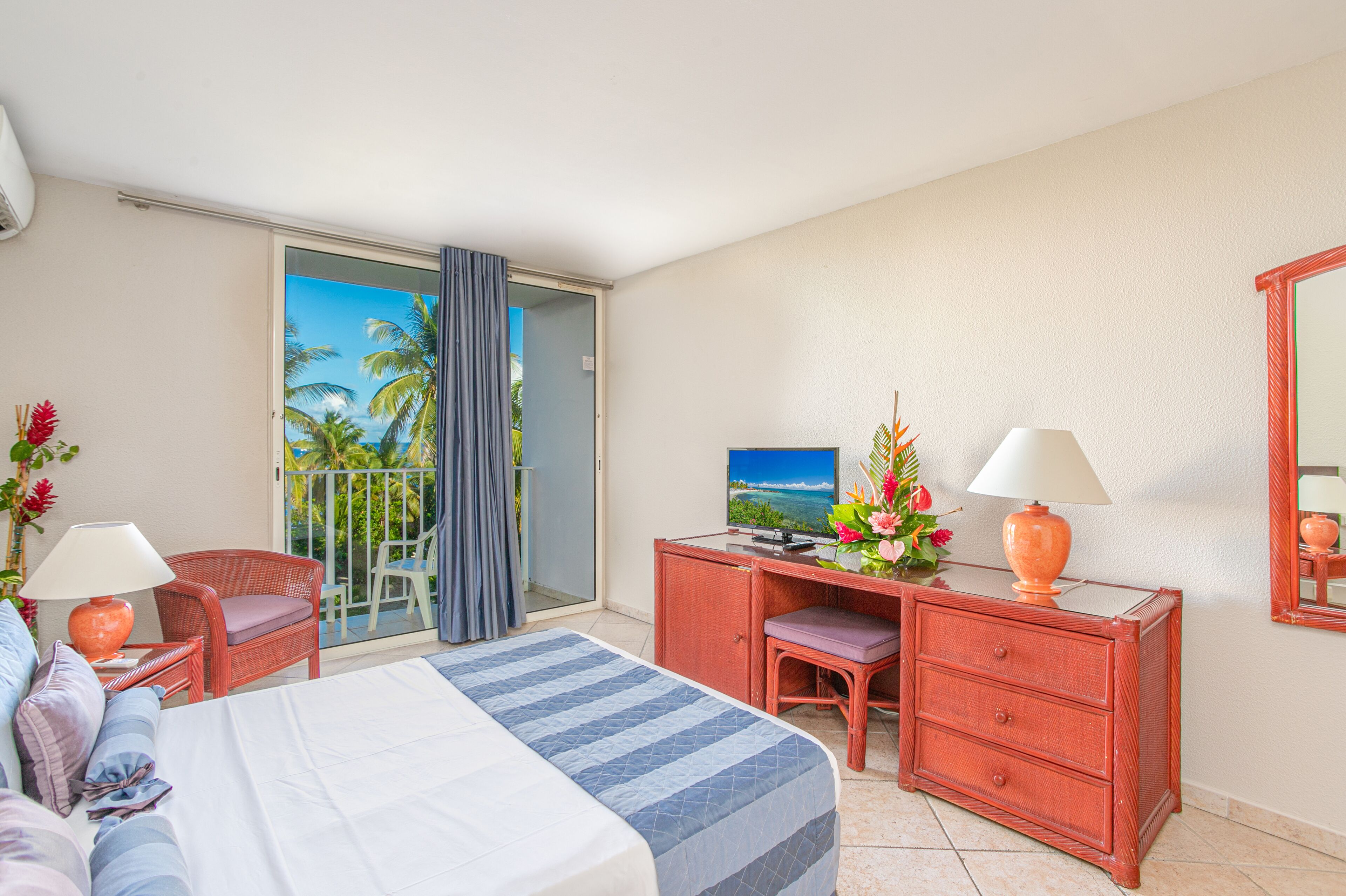 Hotelkamer met een bed, een bureau met een computer, een stoel, een dressoir en een balkon met uitzicht op palmbomen.
