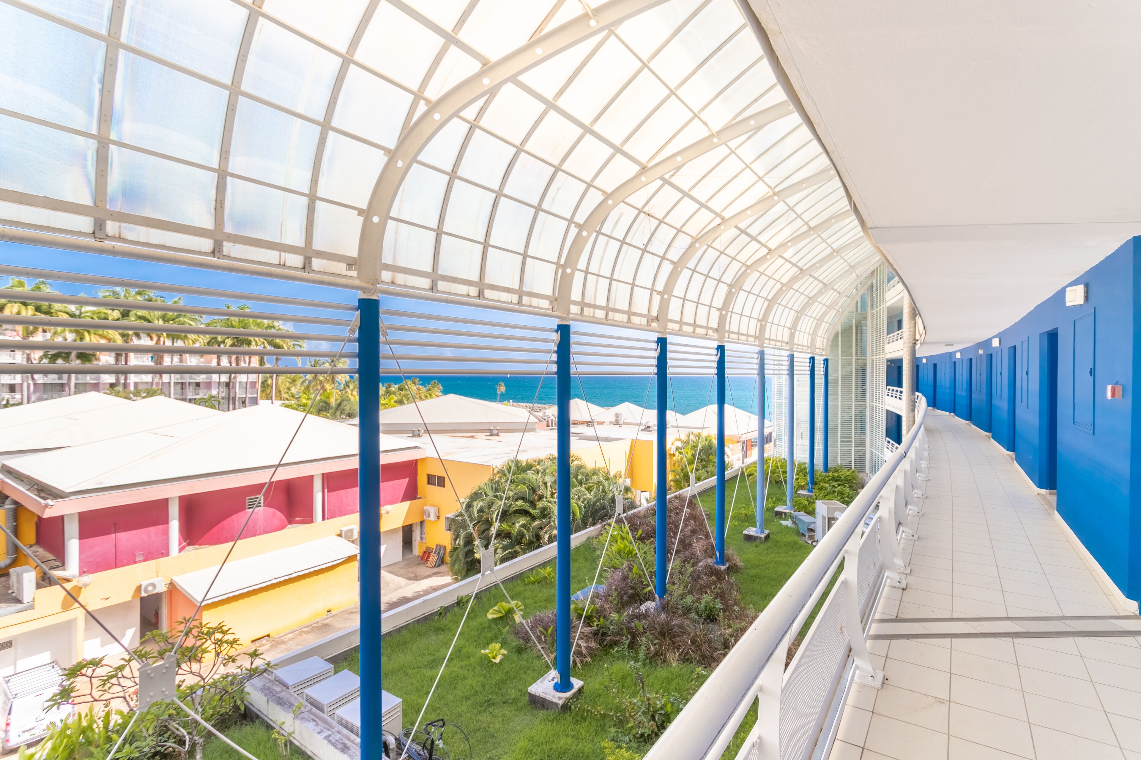A covered outdoor area with a glass ceiling, blue railings, and a view of buildings and greenery below.