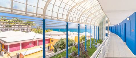 A covered outdoor area with a glass ceiling, blue railings, and a view of buildings and greenery below.