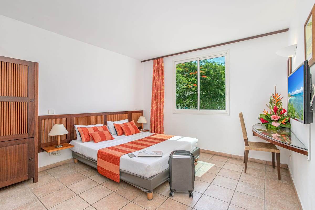 Een hotelkamer met een bed, bureau, stoel, televisie en een raam met uitzicht op bomen.