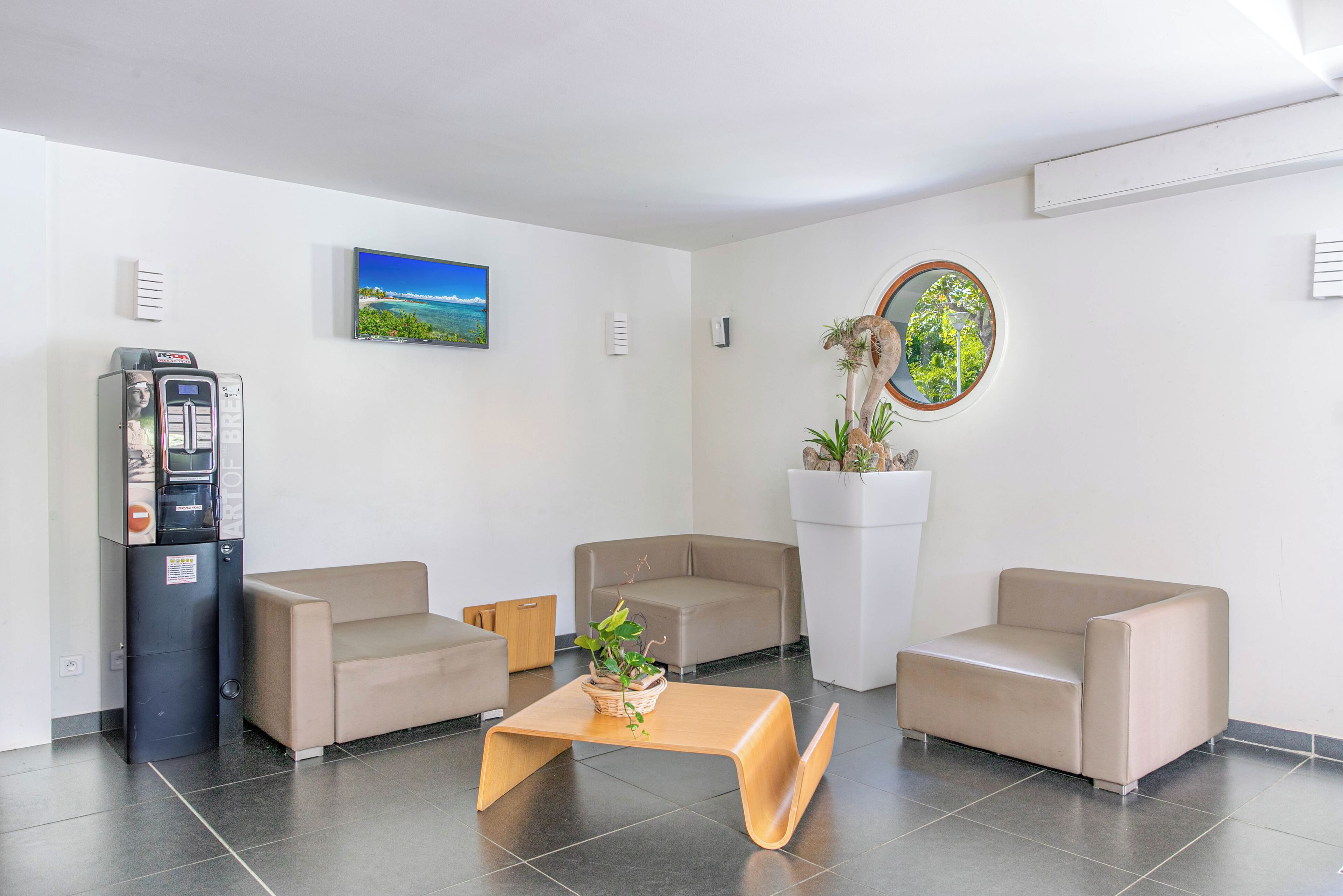 Un área de espera moderna con dos sofás beige, una mesa de centro de madera, una máquina expendedora, un televisor y una planta en maceta.