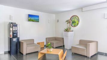 Un área de espera moderna con dos sofás beige, una mesa de centro de madera, una máquina expendedora, un televisor y una planta en maceta.