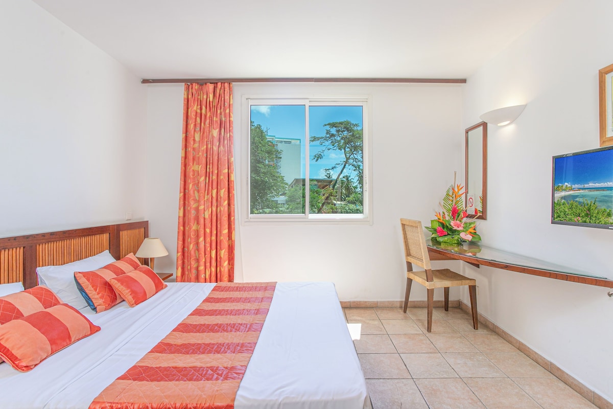 Ein Hotelzimmer mit Bett, Schreibtisch, Stuhl, Fenster mit Blick auf Grünflächen und einem wandmontierten Fernseher.