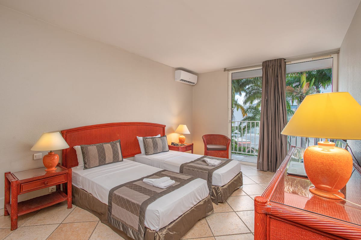 Hotelkamer met twee bedden, een balkon met gordijnen, een rode hoofdbord en een glazen tafel met een lamp.