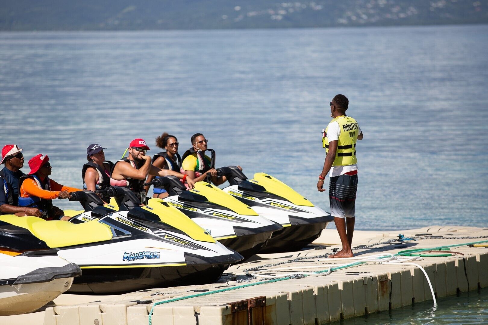 A jet ski rental area with several jet skis lined up on a dock, a person standing beside them, and several people wearing life jackets.