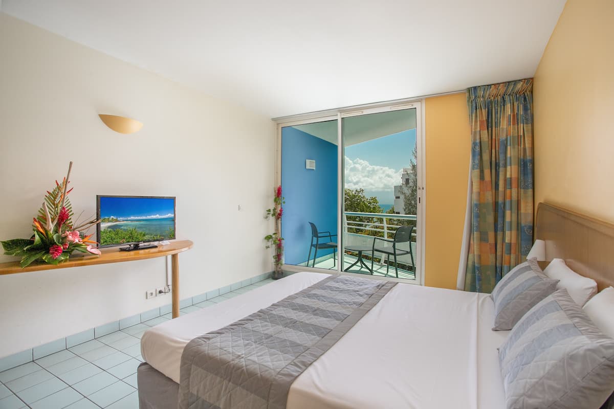 Een hotelkamer met een bed, een televisie, een klein tafeltje met een plant en een balkon met uitzicht op zee.