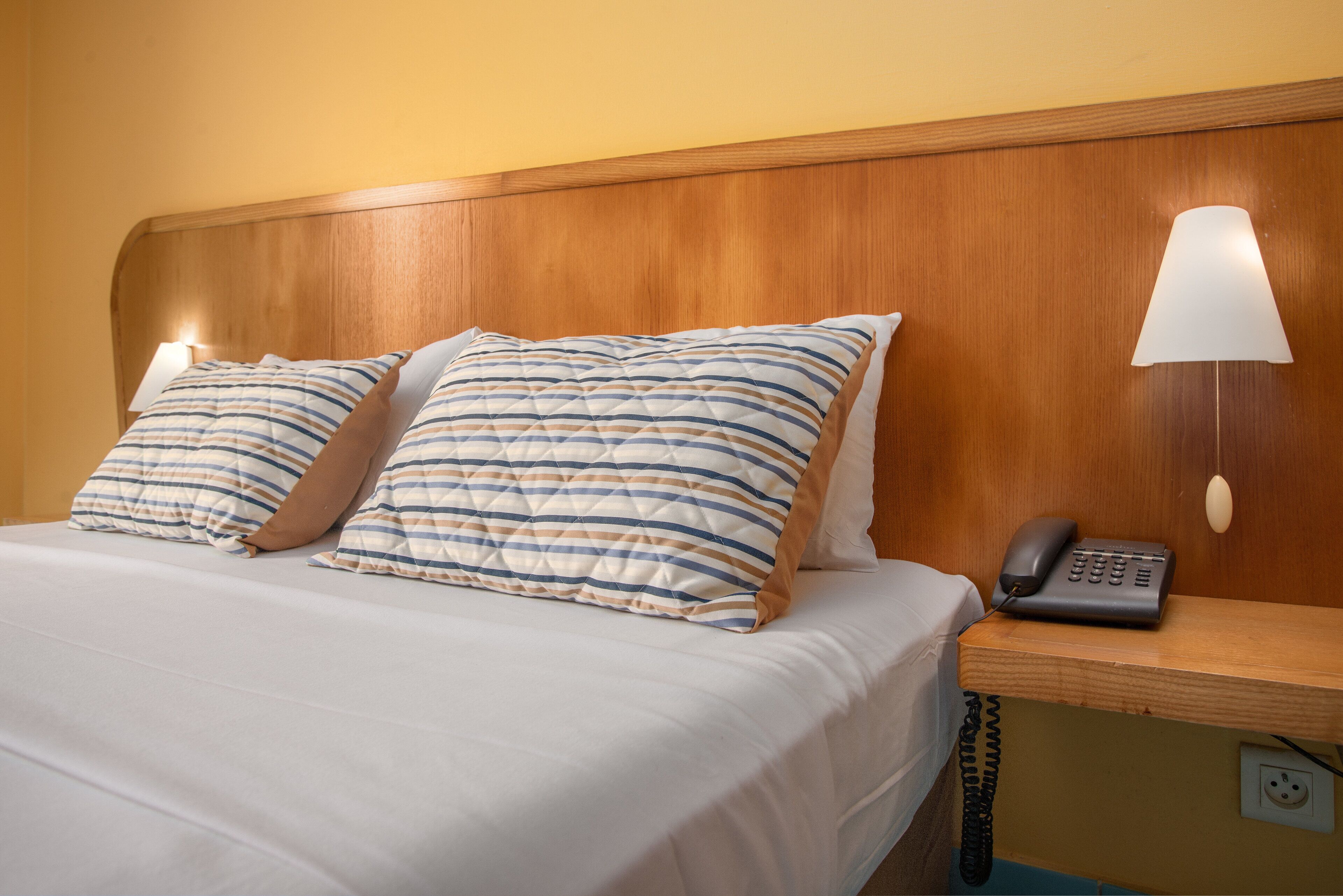 Habitación de hotel con cabecera de madera, dos almohadas a rayas, lámpara de noche y teléfono sobre una mesita de noche de madera.