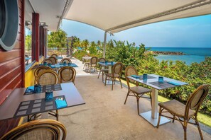 Enjoy al fresco dining with ocean views under a shaded canopy surrounded by lush greenery and palm trees.
