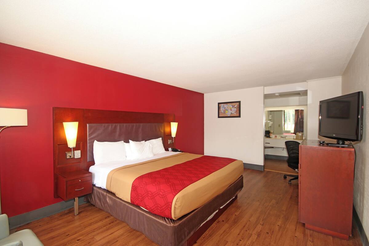 Quarto de hotel com uma cama grande, piso de madeira, parede de destaque vermelha, escrivaninha com cadeira e uma televisão.