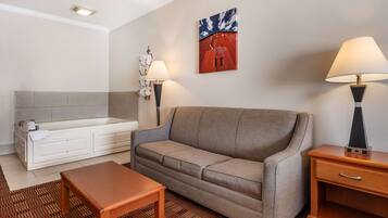 A hotel room with a sofa, a small wooden table, a nightstand with a lamp, a wall-mounted artwork, a bathtub, and a patterned rug.