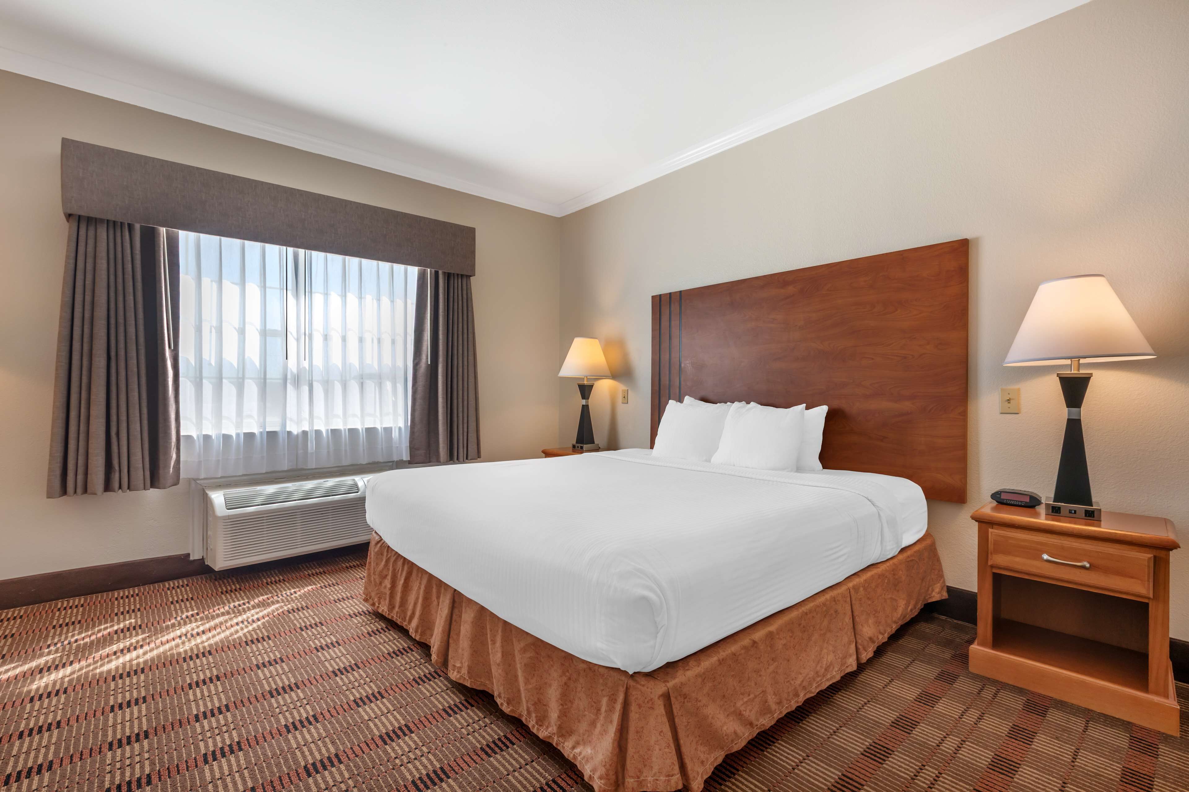 Una camera d'albergo con un letto grande, due lampade ai comodini, una testiera in legno, una finestra con tende e un termosifone.
