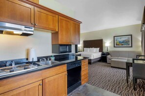A hotel room with a kitchenette, a bed, a sofa, a lamp, and a painting on the wall.