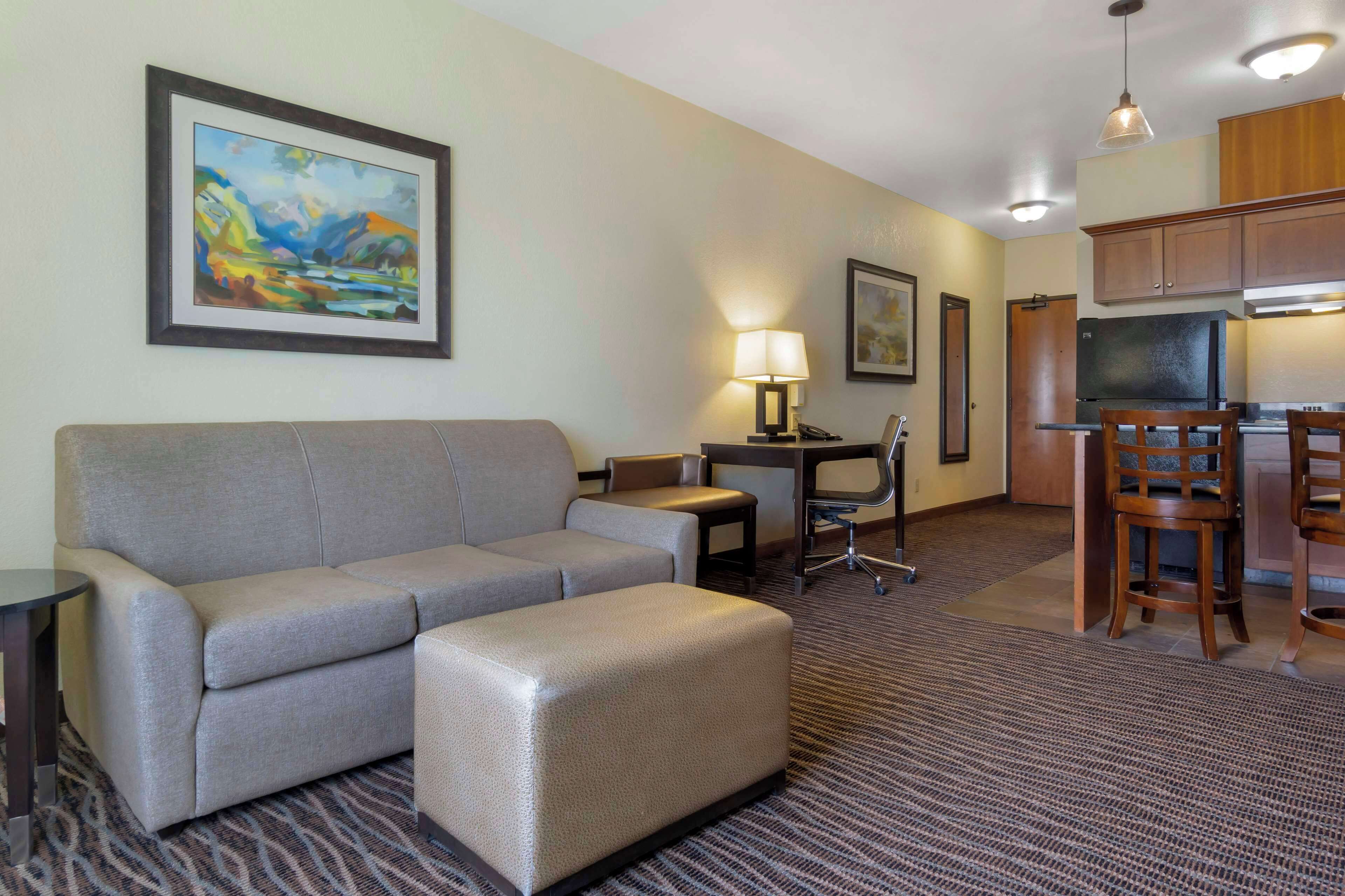 A hotel room with a sofa, ottoman, desk, lamp, and a kitchenette area with bar stools.