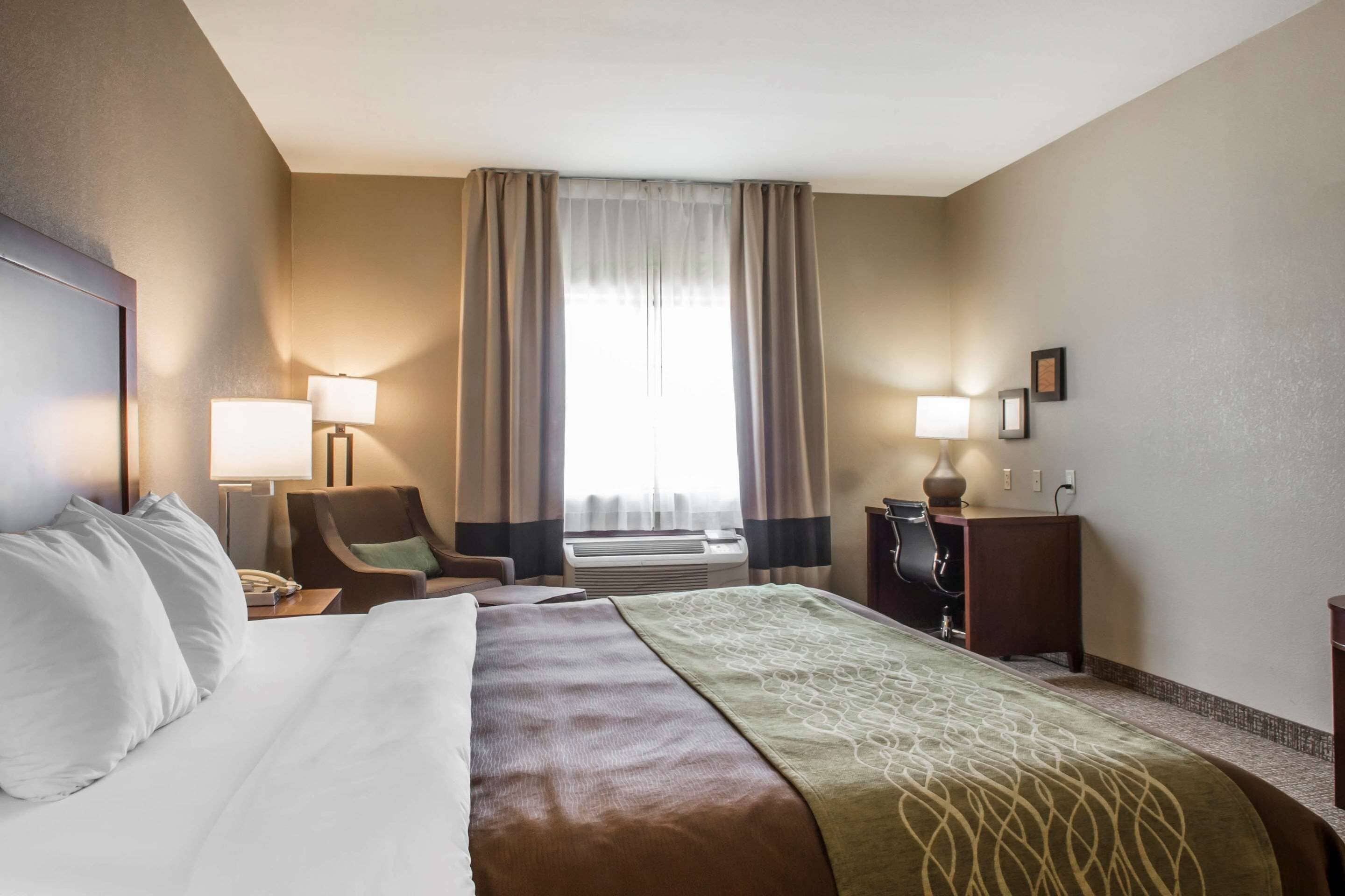 A hotel room with a large bed, a desk, a chair, two lamps, and a window with curtains.