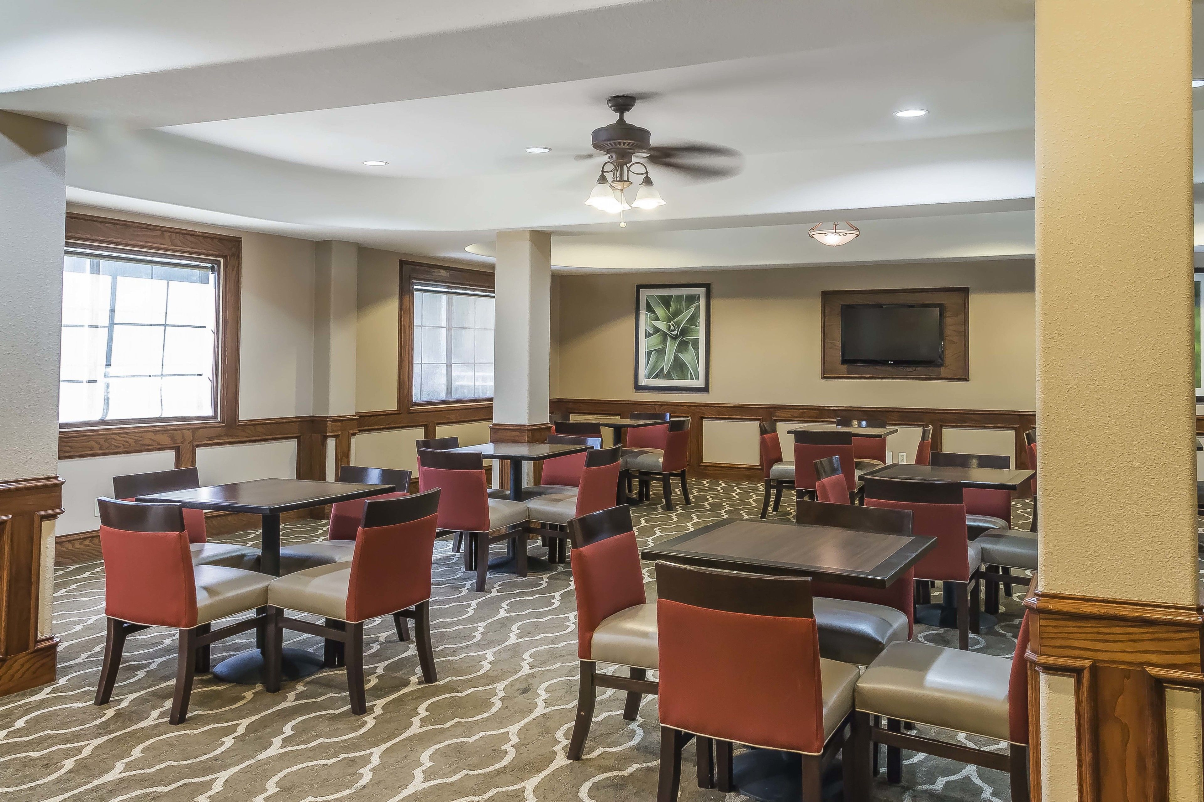 Une salle à manger d’hôtel avec plusieurs tables et chaises, un ventilateur de plafond et un téléviseur fixé au mur.