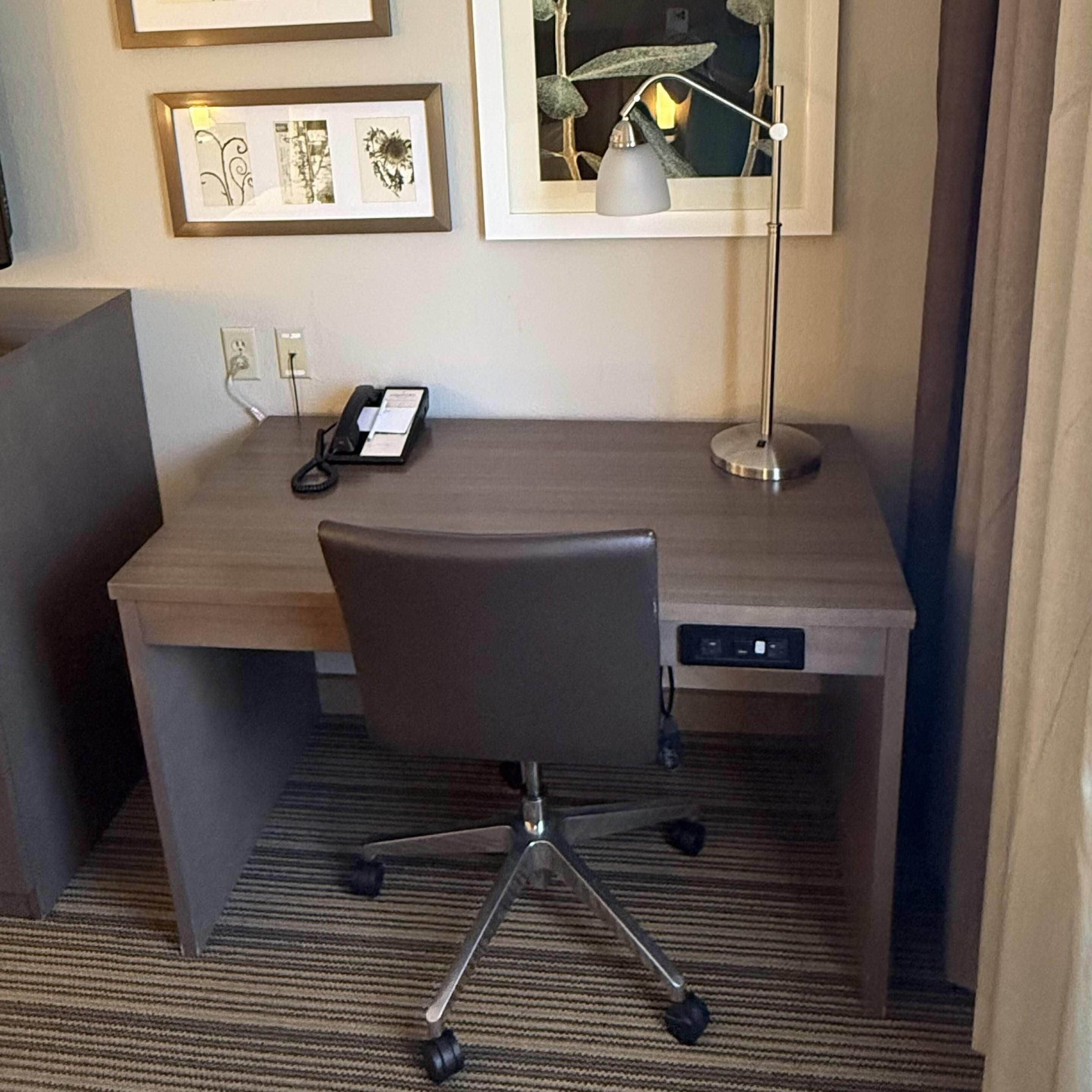 Een hotelkamer met een bureau, stoel, lamp, telefoon en ingelijste foto’s aan de muur.