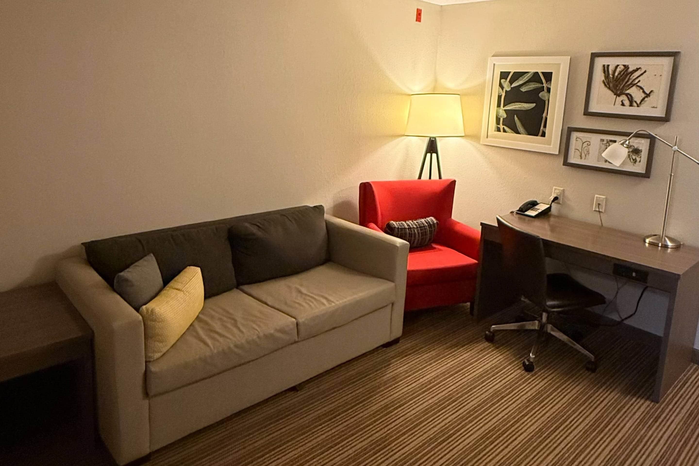 Een hotelkamer met een beige bank, een rode fauteuil, een bureau met stoel, een lamp en botanische kunstwerken in een lijst aan de muur.