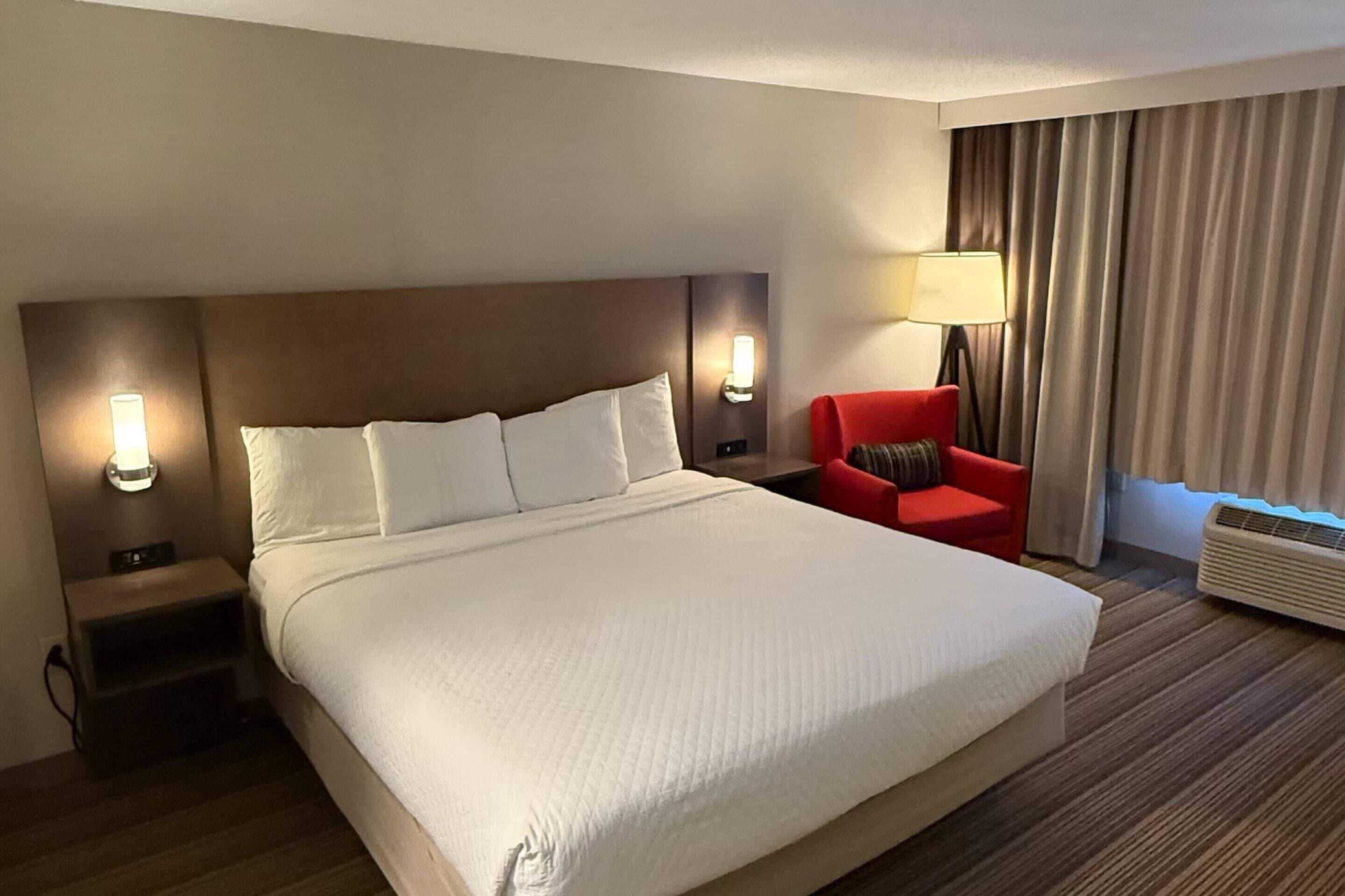 Habitación de hotel con una cama grande, dos mesitas de noche con lámparas, una silla roja, una ventana con cortinas y un equipo de aire acondicionado.