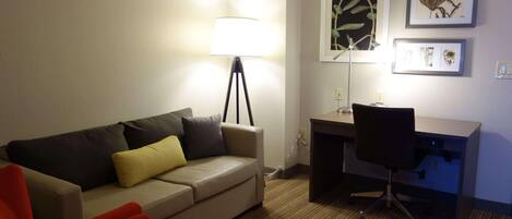 A hotel room with a sofa, a desk, a chair, a lamp, and framed pictures on the wall.