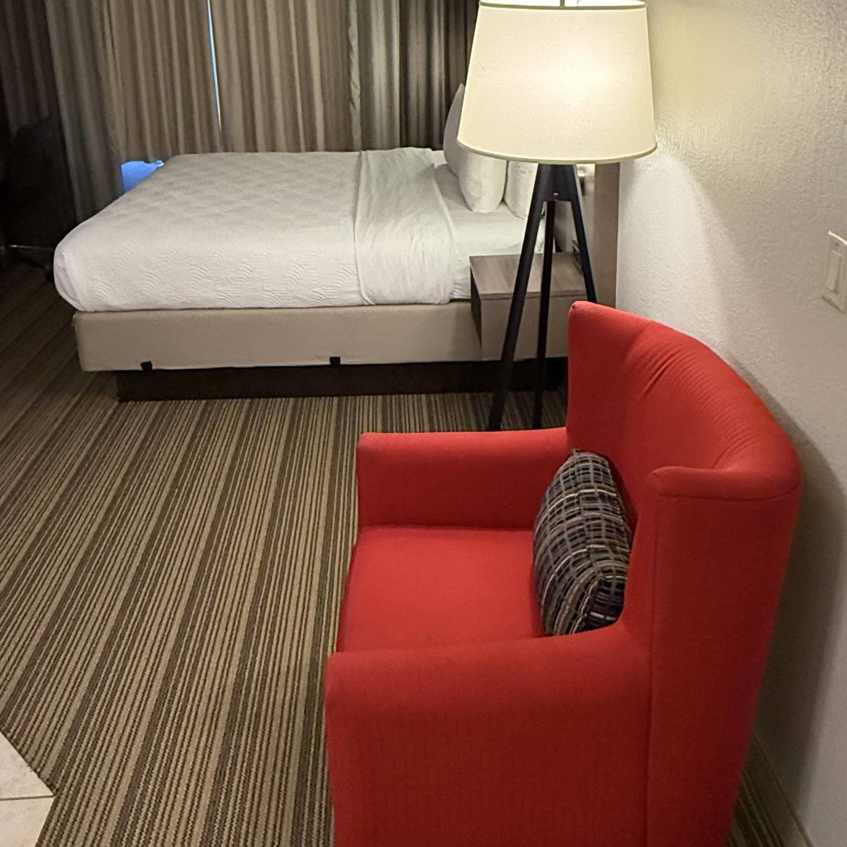 A hotel room with a bed, a red chair, a lamp, and a striped carpet.