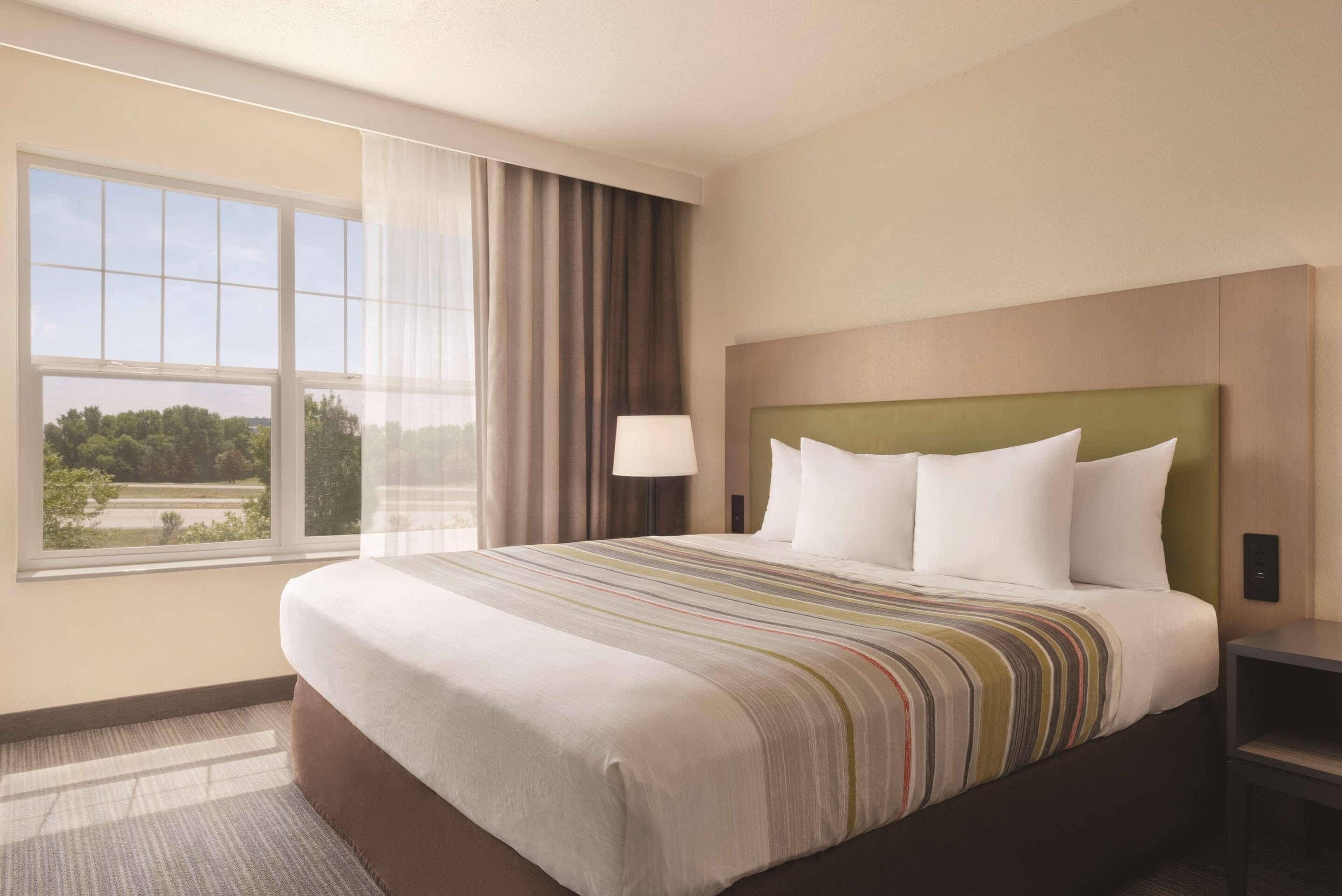 Habitación de hotel con una cama grande, una mesita de noche, una lámpara y vistas al exterior a través de una ventana.