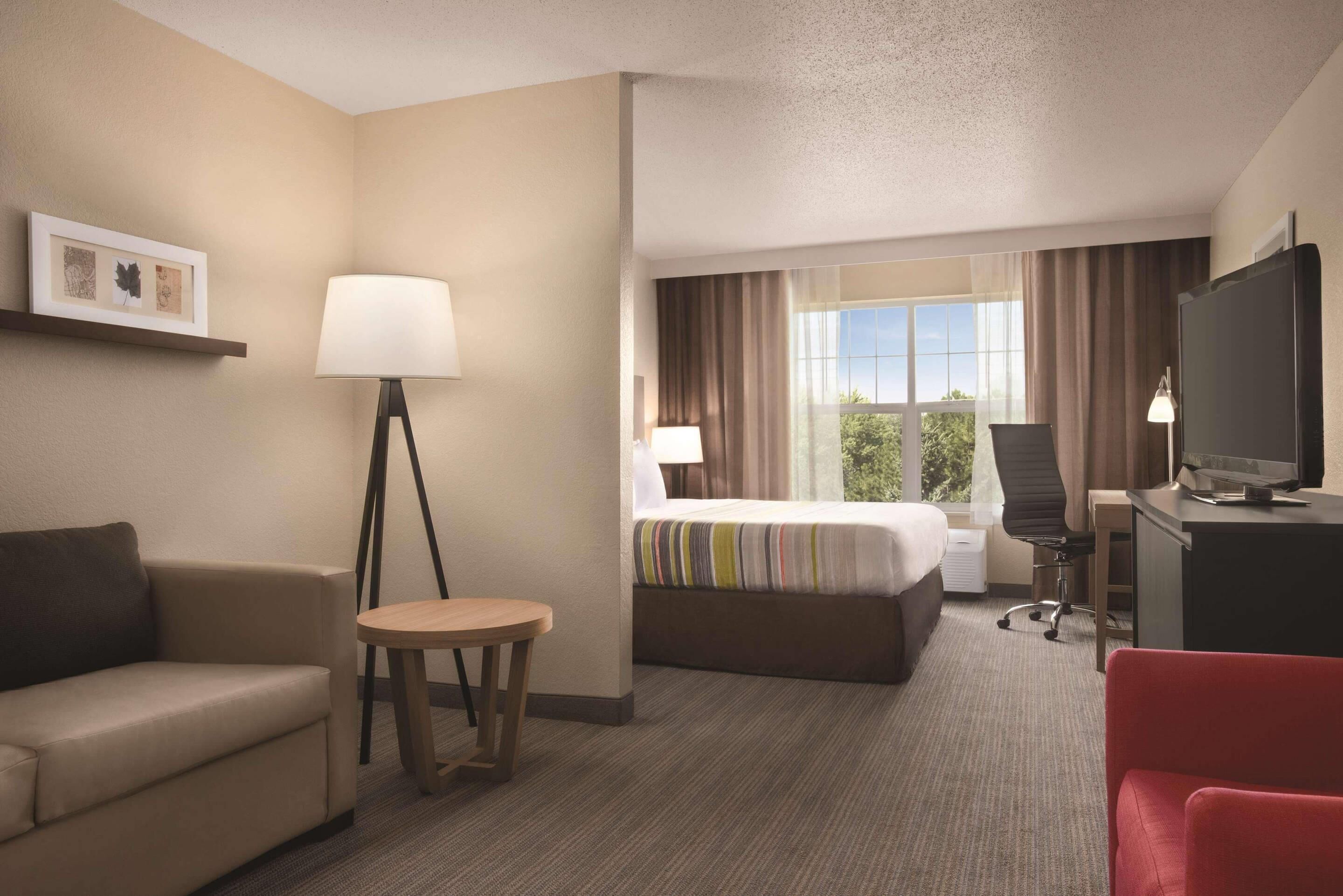 A hotel room with a bed, a desk, a chair, a small table, a sofa, a lamp, and a TV.