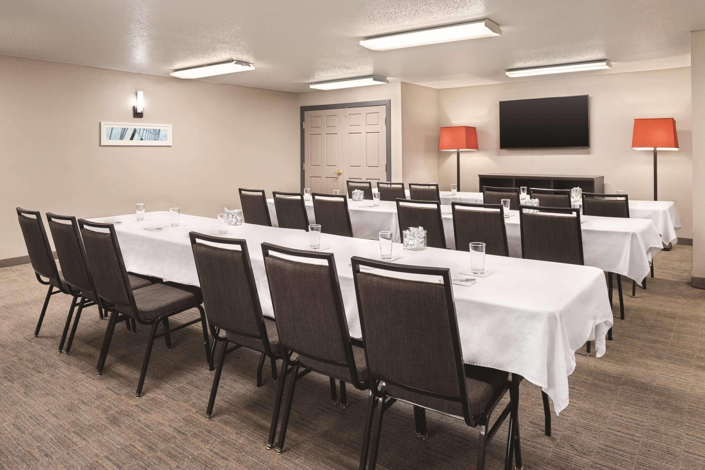 A conference room with multiple tables and chairs, a door, a television, and two lamps.