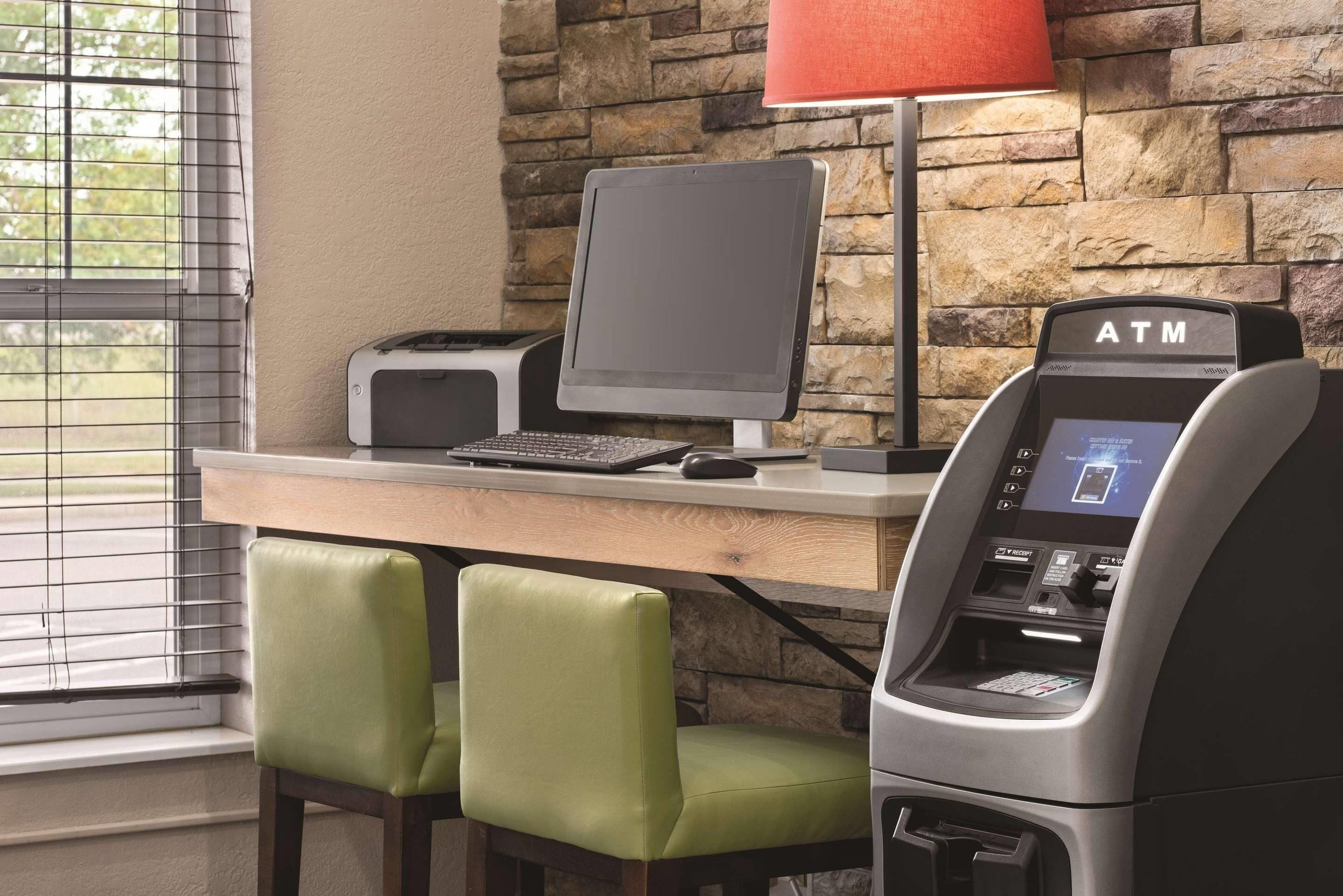 A desk with a computer, an ATM, and two green chairs.