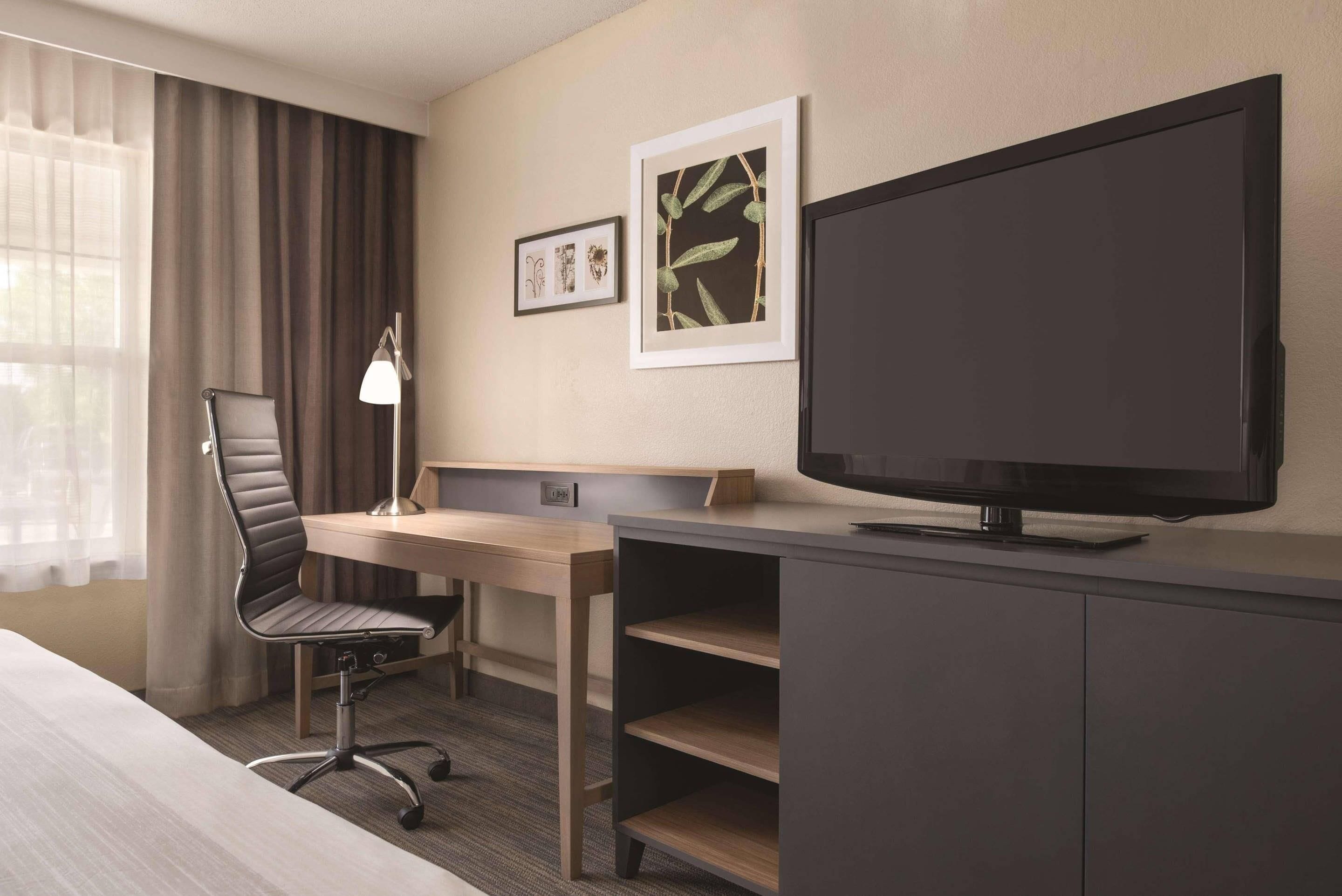Quarto de hotel com cama, escrivaninha, cadeira, televisão e obras de arte na parede.