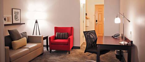 Habitación de hotel con sofá de cuero marrón, un sillón rojo, una silla de oficina negra, un escritorio con lámpara y un teléfono.