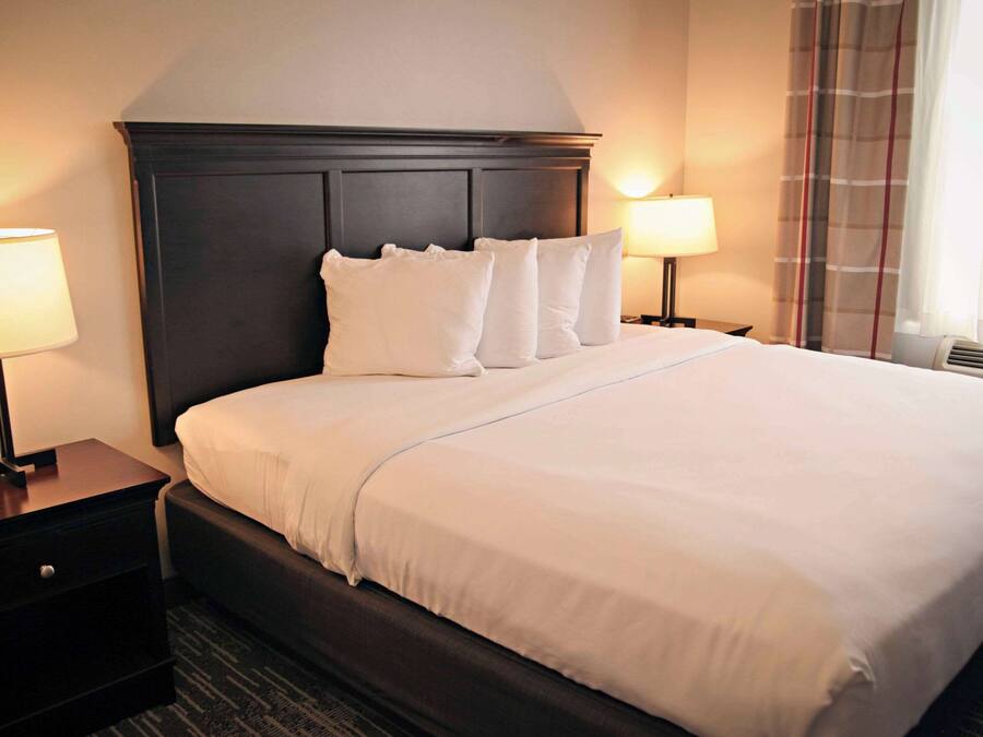 Habitación de hotel con una cama grande, cabecera oscura, dos mesitas de noche con lámparas y una ventana con cortinas.